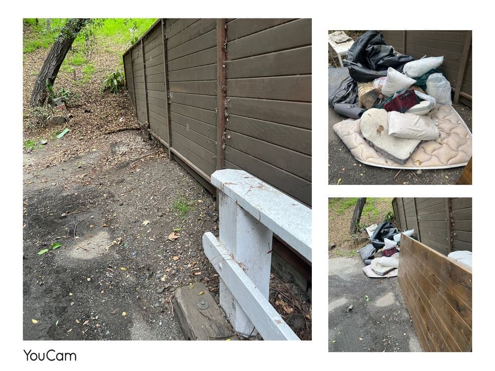 Cluttered outdoor area with discarded pillows and materials beside a wooden fence.